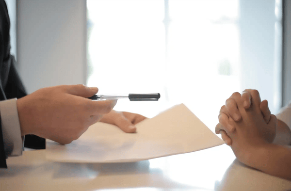 Two people sitting on a table and a person is handing over a paper and a pen to the other person