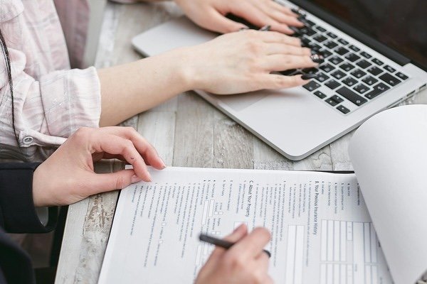 A person typing on a laptop and another person writing on a paperwork