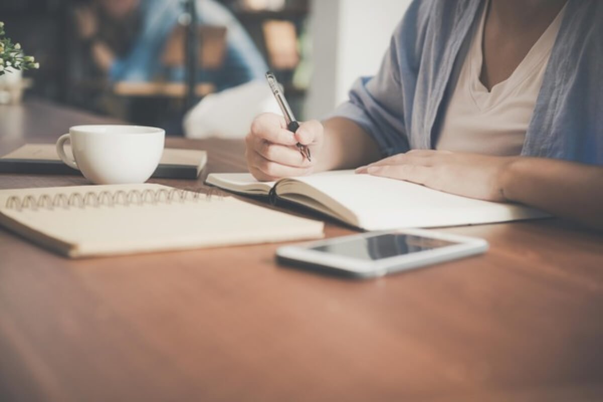 Person writing in a notebook at a café, illustrating budgeting, financial planning and practical money lessons for everyday life.