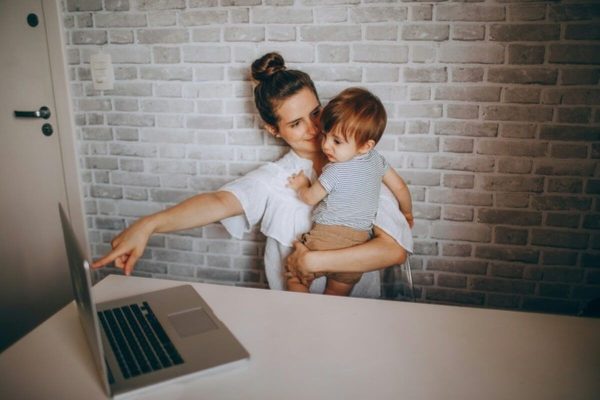 Mother working on a laptop while holding her child, highlighting policies that support working mothers balancing career and caregiving.