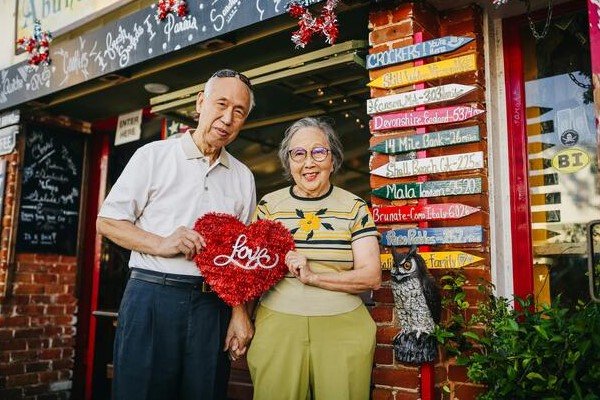 A photo of an elderly couple holding a red heart-shaped pillow with the word love