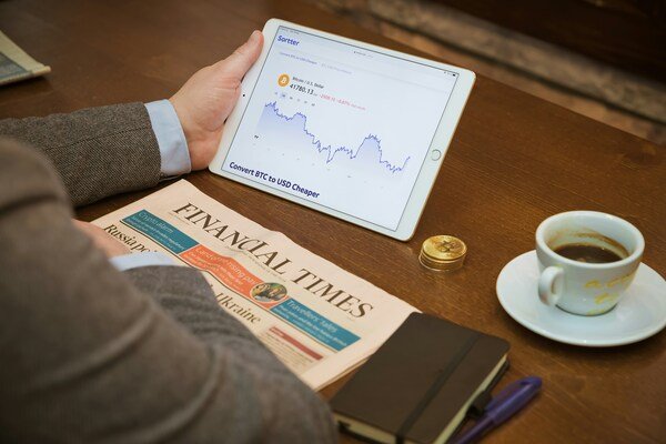 A photo of a person's hand rests on a table, holding a gadget placed beside a newspaper and a cup of coffee