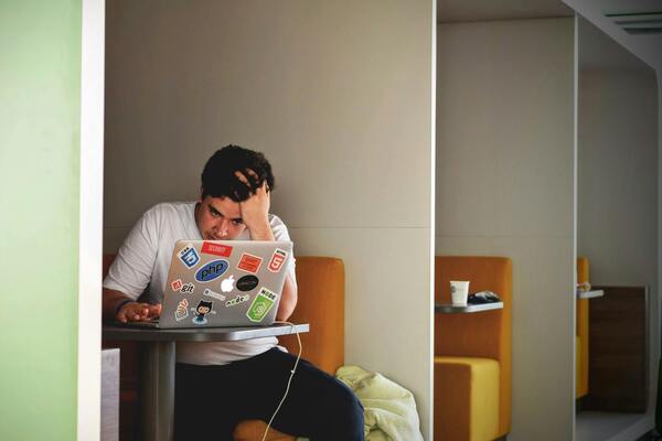 A photo of a man wearing a white shirt while using a laptop, looking frustrated