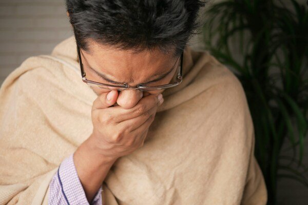 A man wearing eyeglasses with a brown blanket draped over his shoulders holds his nose, appearing to be unwell