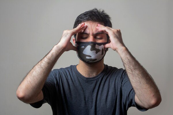 A photo of a man wearing a face mask holds his head with both hands, appearing to be in pain or discomfort