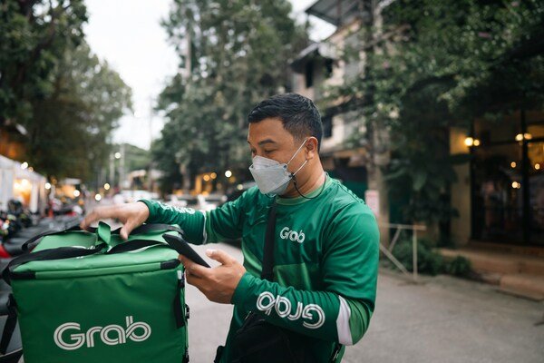A delivery worker wearing a face mask and green jacket, holding a phone with one hand and a large green insulated Grab delivery bag on the motorbike in a street setting.