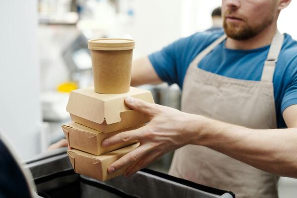 A person wearing an apron and blue shirt holds a stack of eco-friendly cardboard takeaway containers in a bright kitchen or service-area setting.