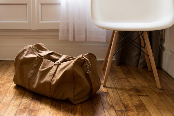 A brown duffel bag resting on a wood-tone floor next to a white-and-wooden chair with angled legs.