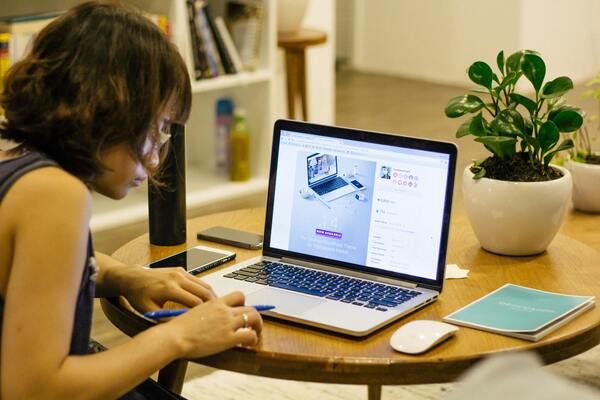 A woman in a black tank top uses a laptop at a round wooden table.