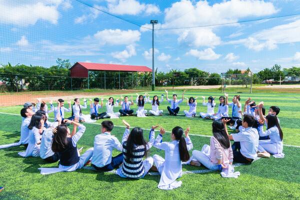 A group of people sit on grass raising their hands outdoors in daylight.