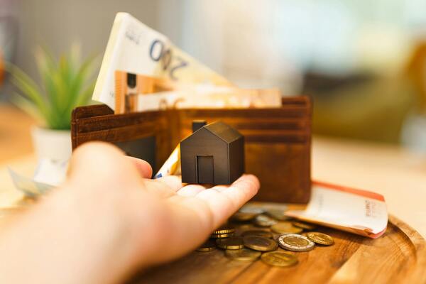 A miniature house in a person's palm with money in the background