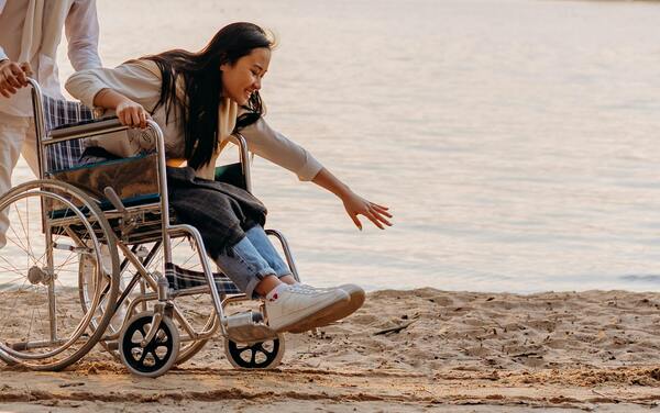 Woman in a wheelchair reaching toward the sea at a sandy beach, symbolizing accessibility, inclusion, and quality of life.