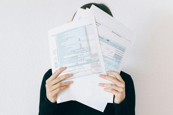 Person holding multiple financial forms covering their face, representing paperwork stress, financial obligations, and administrative challenges.