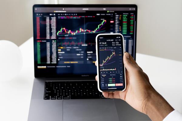 Person holding smartphone with stock market charts in front of a laptop, representing investing, trading, and financial risk management.
