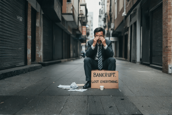 Distressed businessman sitting on an empty street holding a bankrupt lost everything sign, representing financial failure and bankruptcy impact