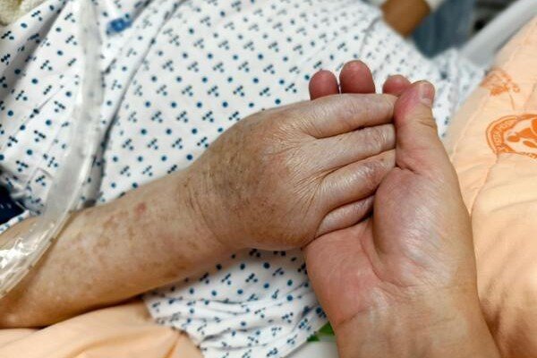 “Close-up of caregiver holding elderly patient’s hand in hospital bed, symbolizing compassion, support, and palliative care
