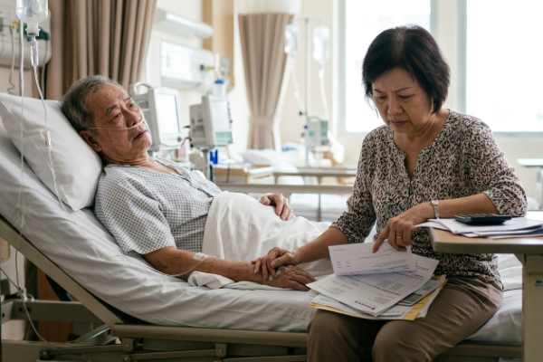 An Asian woman sits beside an elderly man in a hospital bed, offering support and comfort during his recovery.