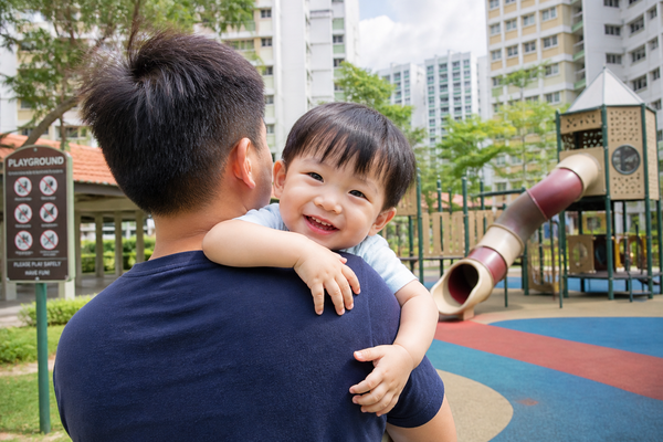 A man holds a child in front of a colorful playground, surrounded by swings and slides on a sunny day.